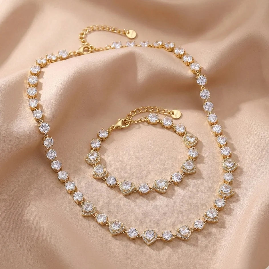 Podroben pogled na lesketajočo zlato zapestnico iz vrste srčastih členov s kamni, kot jo nosi model. Zapestnica je oblikovana tako, da tvori popoln set z biserno ogrlico z zlato pentljo in srčkom.