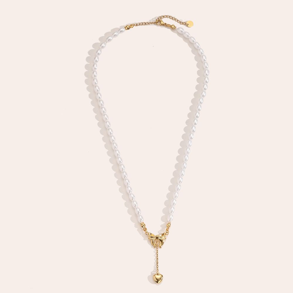 Eleganten darilni set, primeren za dan žena, položen na bež svilo. Prikazuje biserno ogrlico z detajlom zlate pentlje in visečim srčkom ter par klasičnih bisernih uhanov.