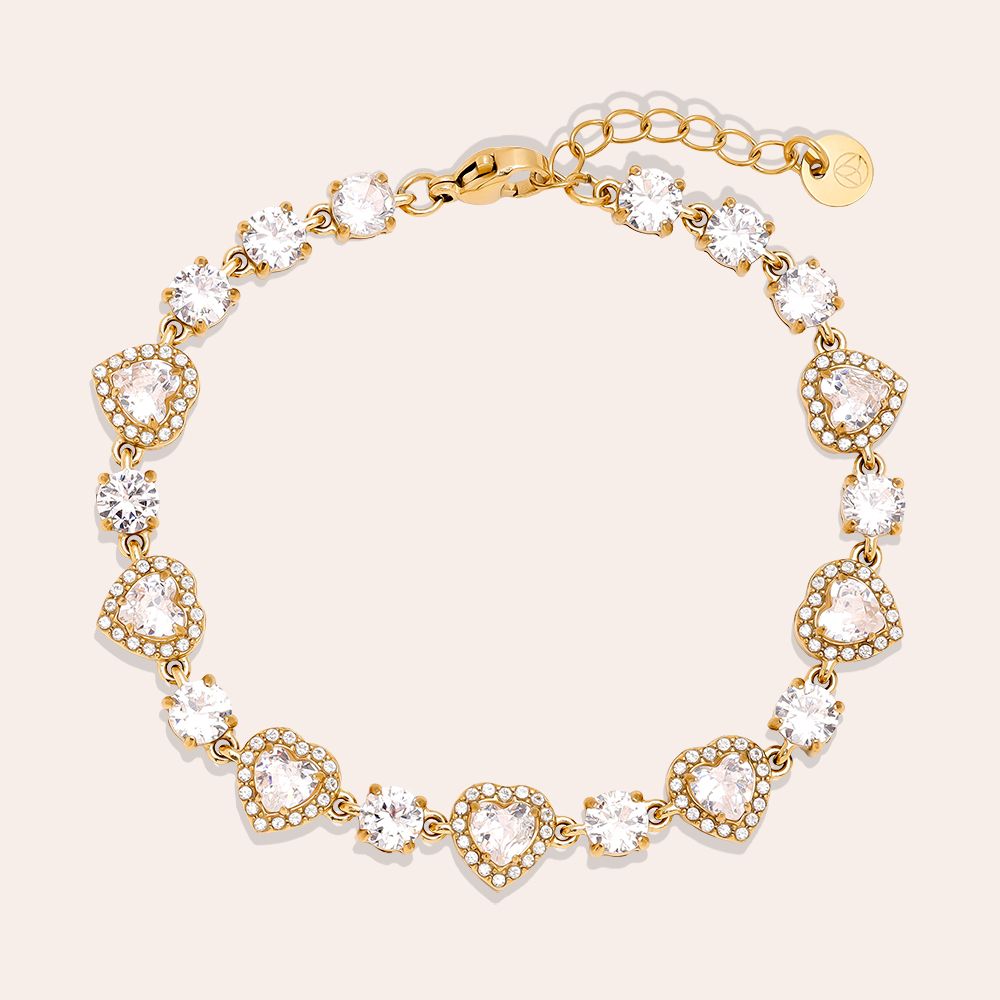 Ženska nosi elegantno zapestnico z zlatimi srčki in cirkoni. Ta modni dodatek je del čudovitega darilnega seta, ki ga dopolnjuje biserna ogrlica z obeskom pentlje, idealno za posebne priložnosti.